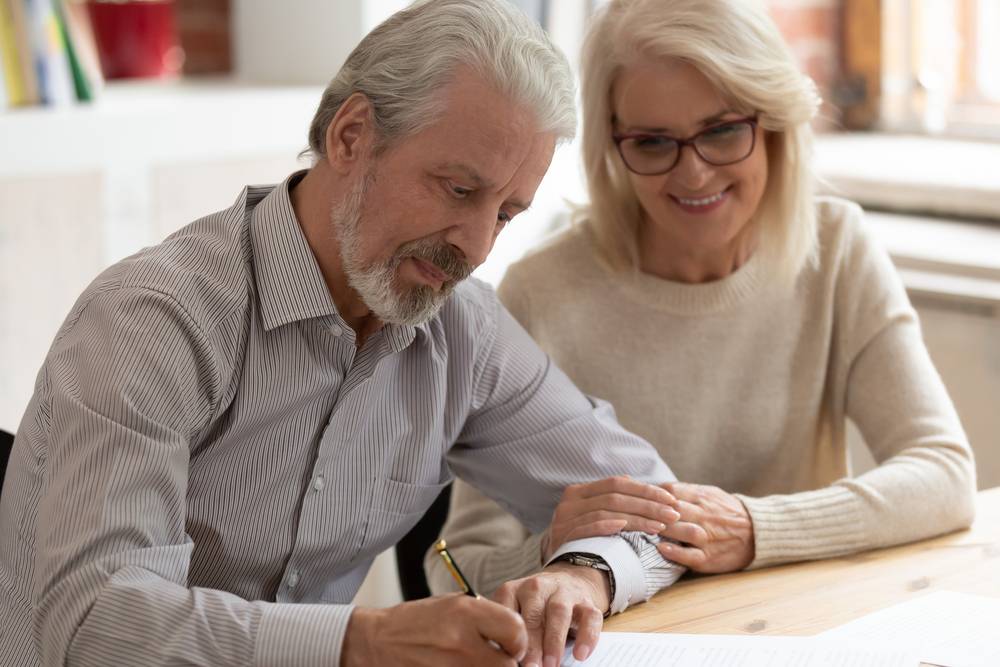 Older man signs documents at a table while a smiling woman sits beside him, hand on his arm.