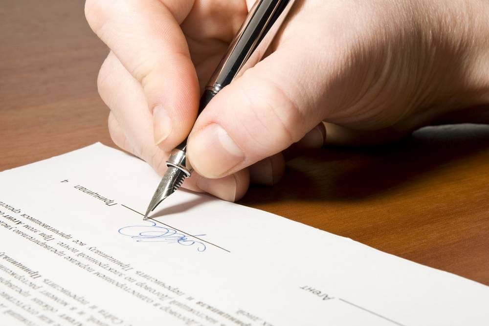 A hand holding a pen signing a document on a wooden desk.