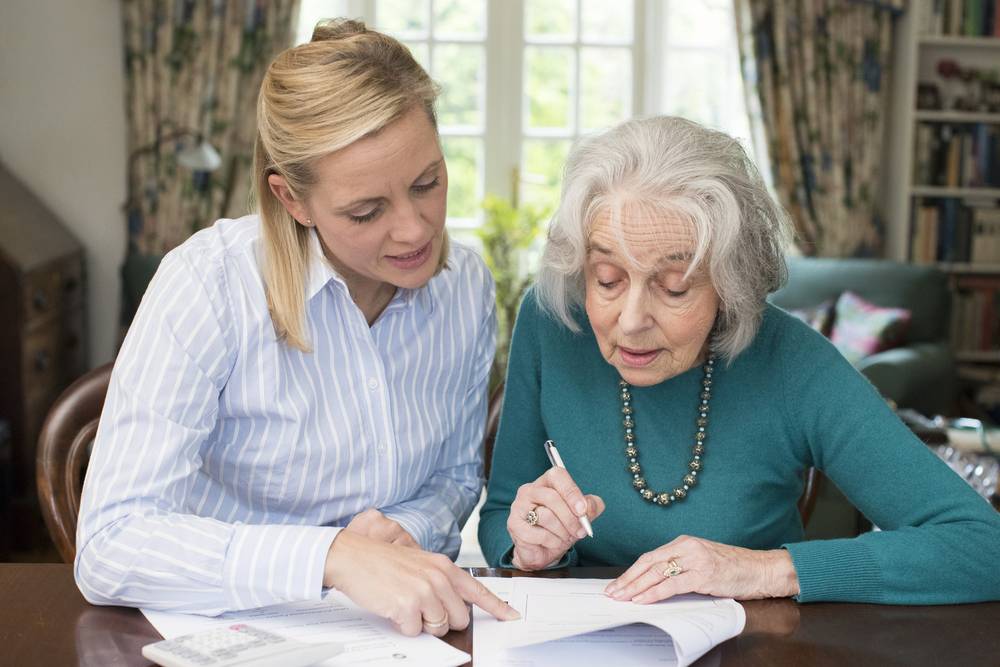 A younger woman helps an older woman review and fill out paperwork at a table indoors.
