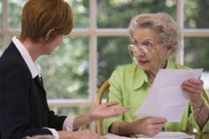 Elderly woman holding paperwork talks to another woman at a table with a window in the background.